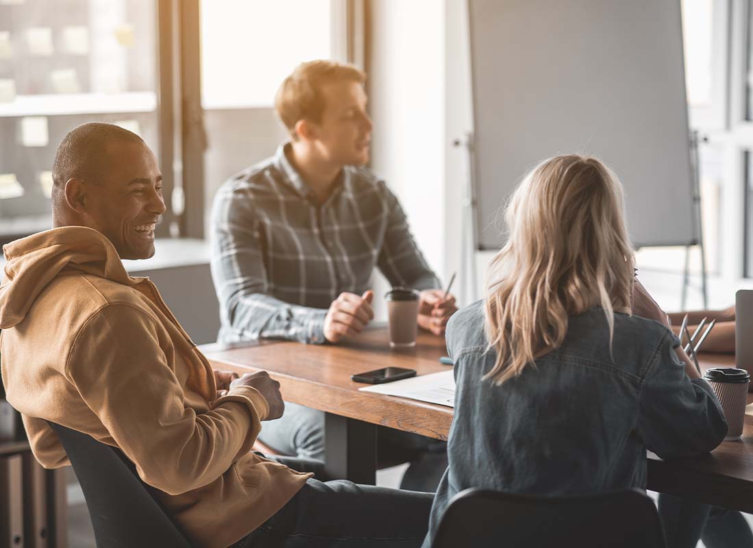 Group Voluntary Insurance - Four Young People Sitting at an Office Conference Table Together and Conversing and Laughing with Coffee Cups on the Desk